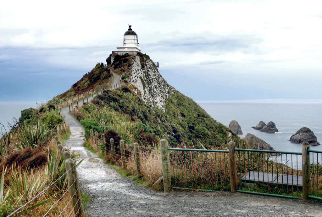 Winding grassy path up rugged hill to solitary lighthouse by ocean with cliffs under overcast sky Wallpaper