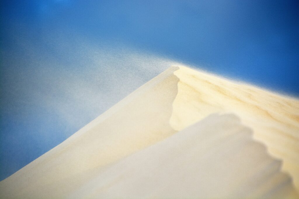 Serene windswept desert dune with sunlit ridges and drifting sand under clear blue sky wallpaper