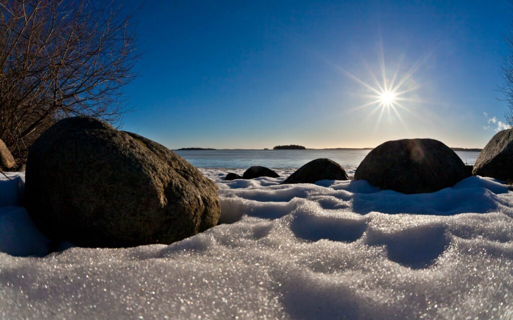 Winter beach wallpaper with large rocks on snow and calm sea under clear blue sky