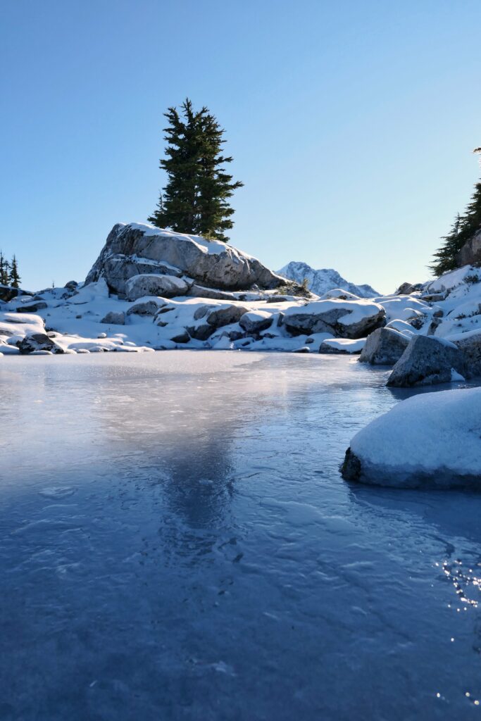 Winter wallpaper of frozen lake with snow-covered rocks and evergreen trees under blue sky