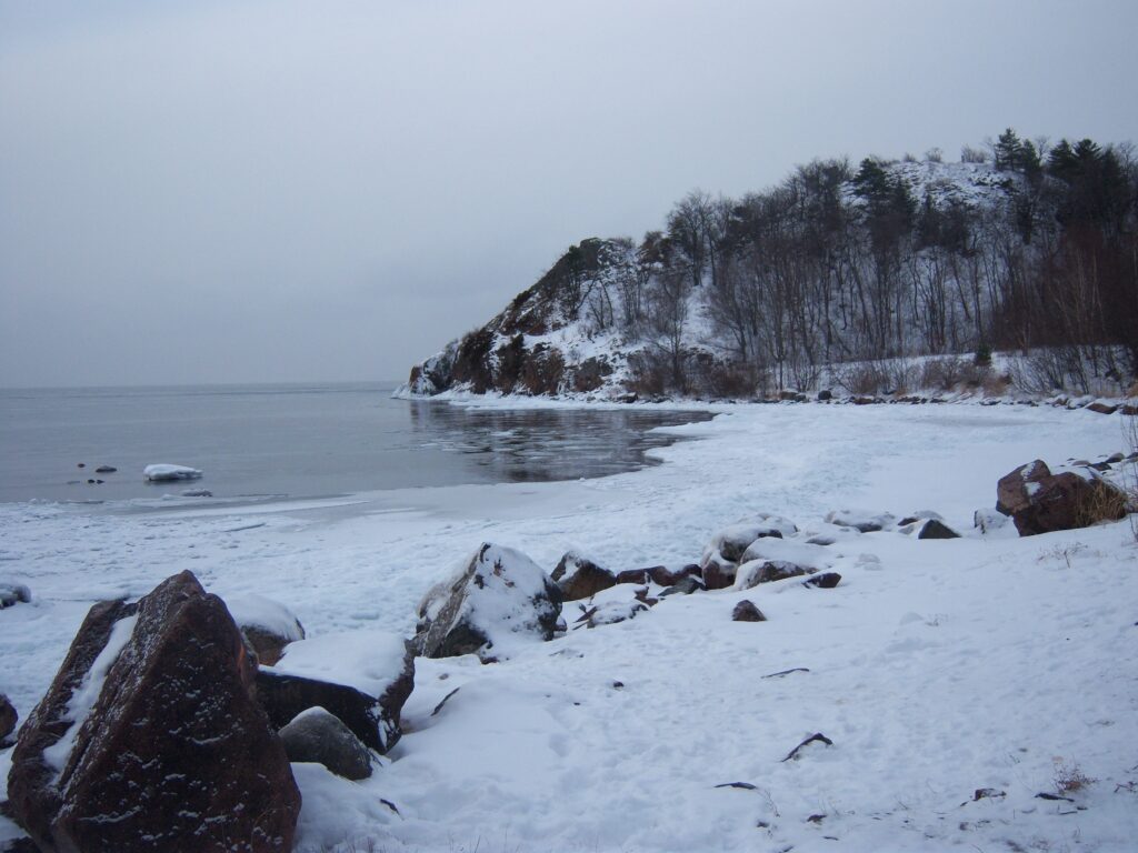 Serene winter landscape with snow-covered rocks and tree-lined lake under overcast sky wallpaper
