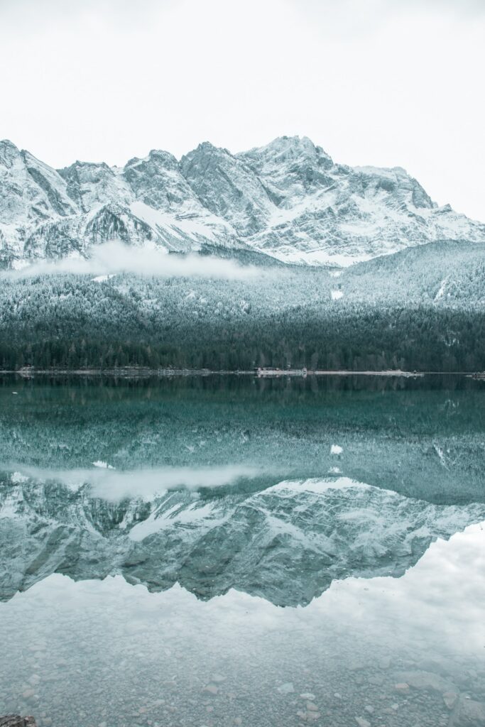 Serene winter lake reflecting snow-covered mountains and dense forest with icy blue water Wallpaper