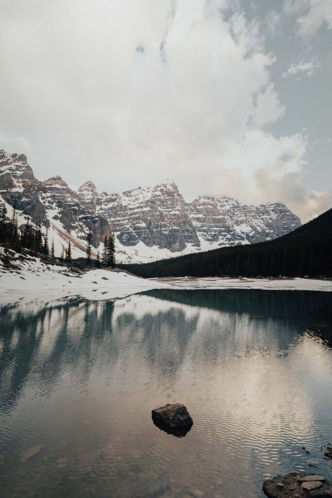 Serene winter lake with snowy stones reflecting majestic mountains and soft clouds background Wallpaper