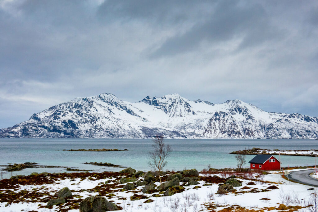 Winter landscape with solitary tree red cabin snowy mountains and calm lake serene scene Wallpaper