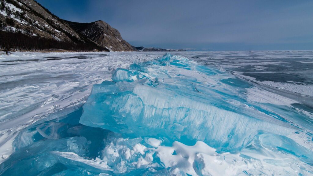 Frozen lake with thick blue ice snowy rocks and distant mountains under gray winter sky wallpaper