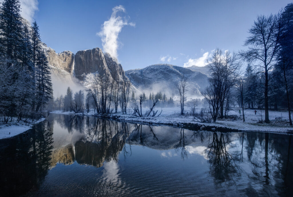 Serene winter river reflecting snow-covered trees and mountains with morning fog under blue sky Wallpaper