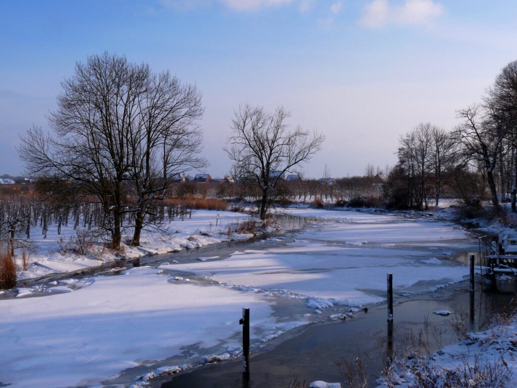 Serene winter landscape with frozen river bare trees snow patches and soft blue sky wallpaper background