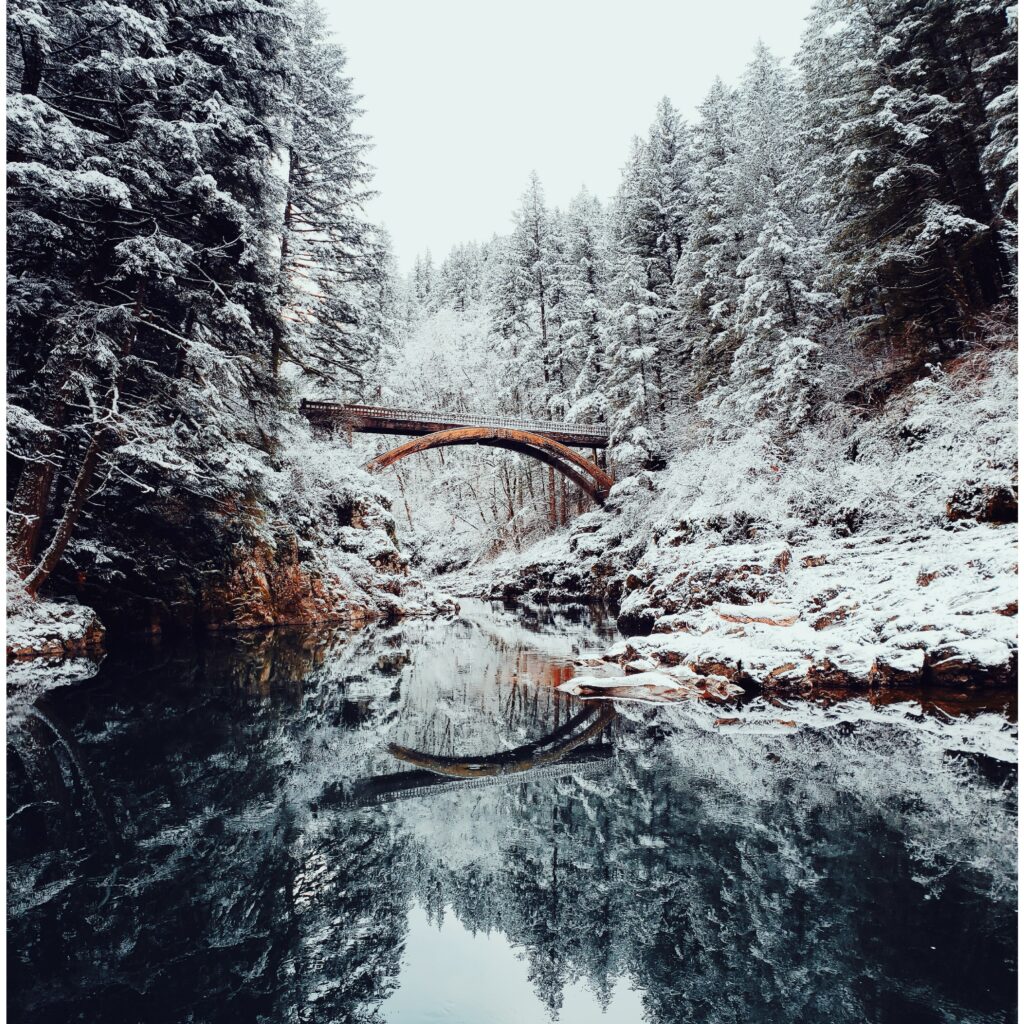 Rustic bridge over calm river surrounded by snow-covered trees reflecting in winter landscape Wallpaper