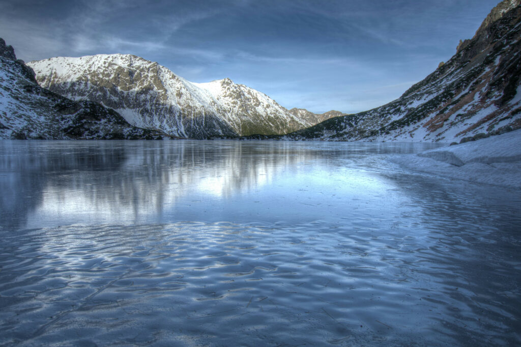 Snow-covered mountains reflected on frozen lake with icy textures and crisp blue hues winter landscape Wallpaper