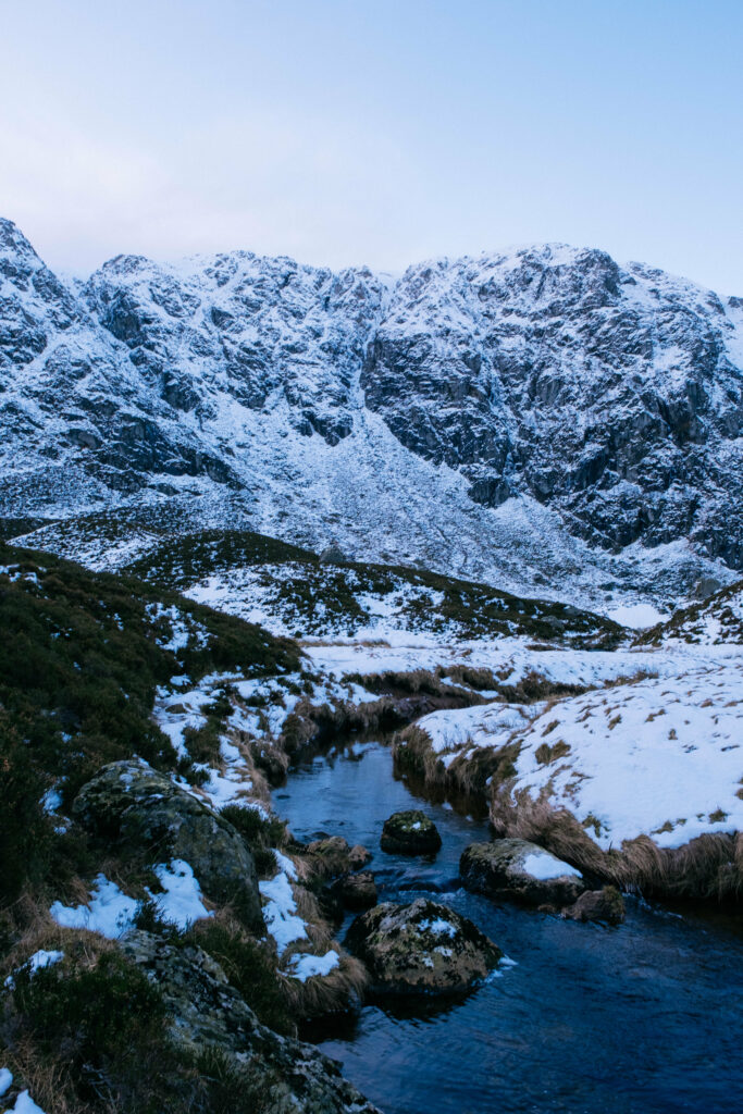 Serene winter landscape with snow-covered mountains gentle stream and grass under blue sky wallpaper