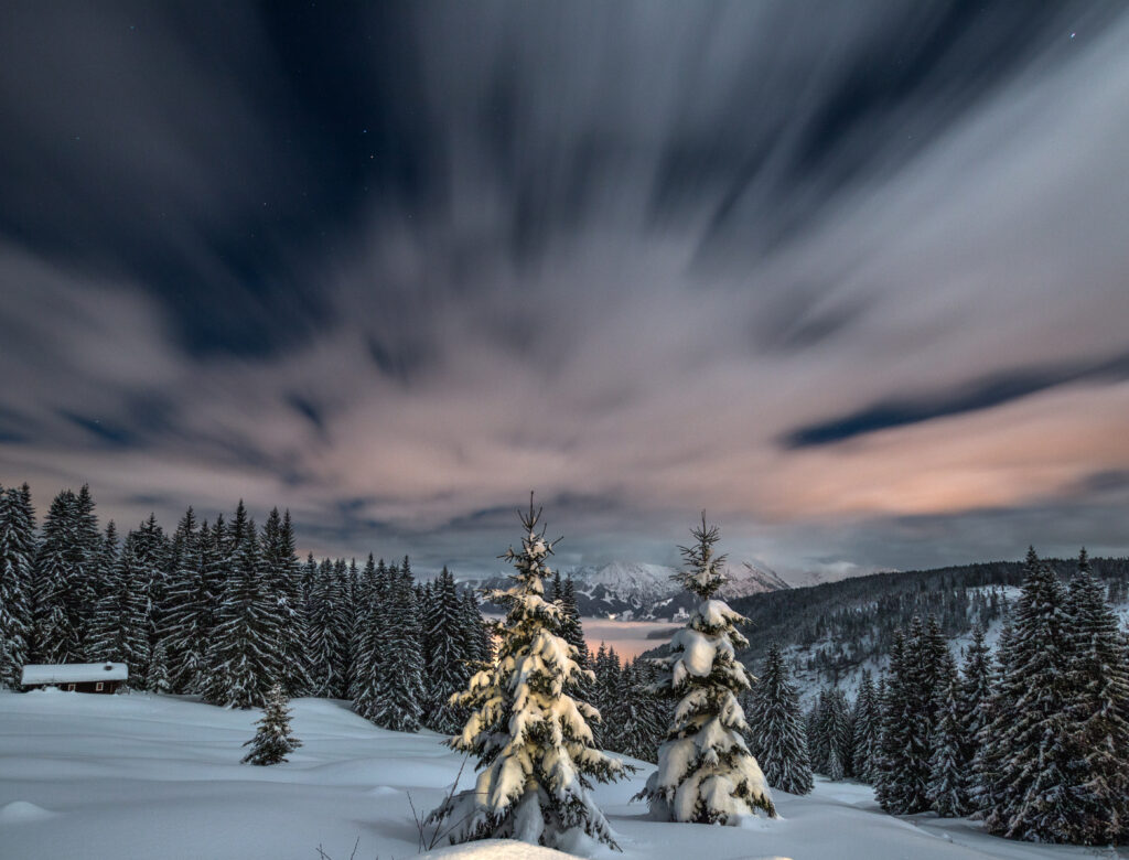 Serene snowy fir forest and mountains under pastel sunset sky with flowing clouds winter landscape Wallpaper