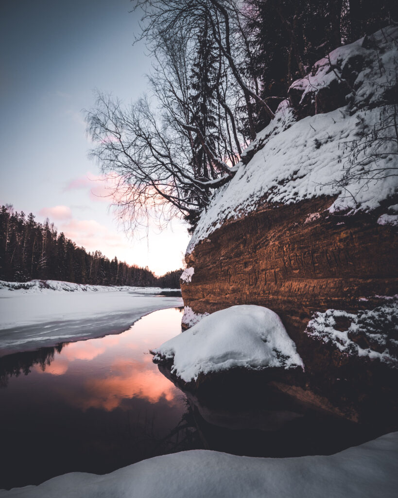 Snowy cliff and calm lake at sunset framed by leafless trees and dense evergreen forest winter wallpaper