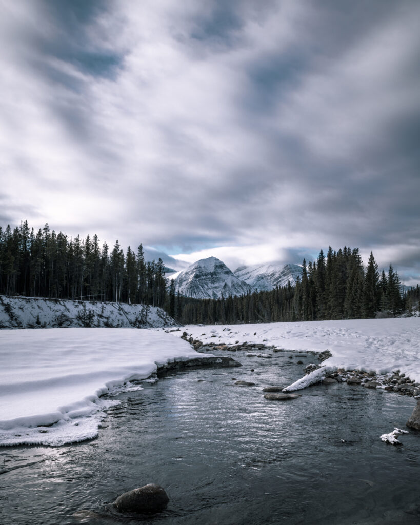 Serene winter landscape with river flowing through snow-covered pine forest and majestic mountains Wallpaper