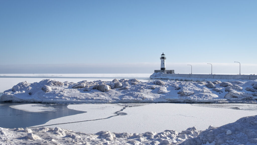 Serene winter landscape with lighthouse on snowy coast ice-covered rocks calm water background Wallpaper
