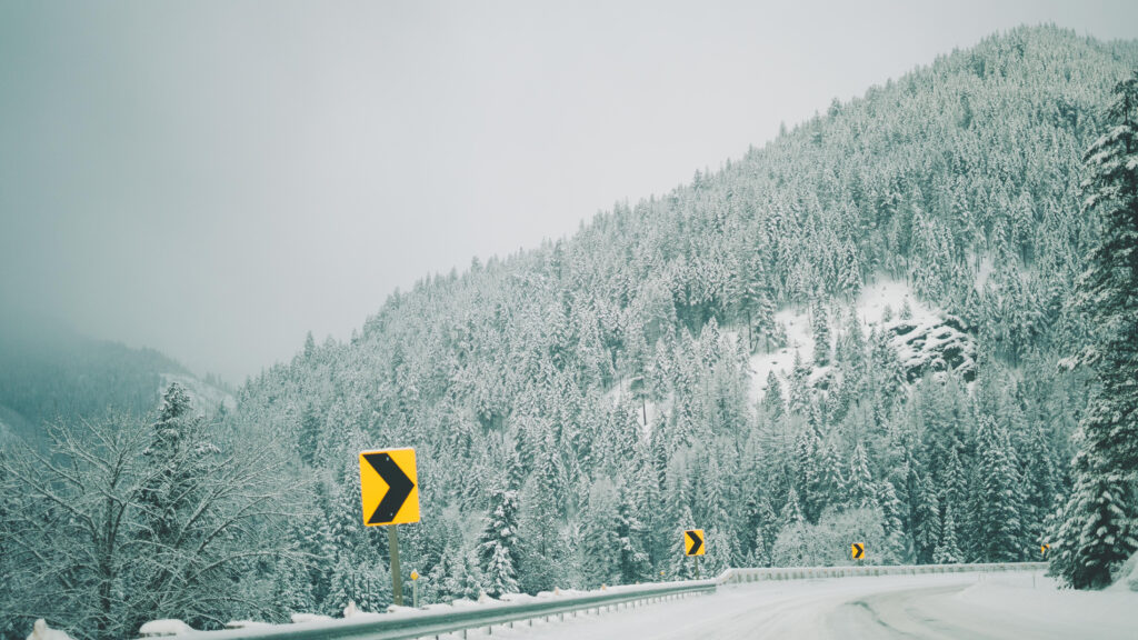 Serene snow-covered mountain forest with bright yellow road signs on winding winter road wallpaper