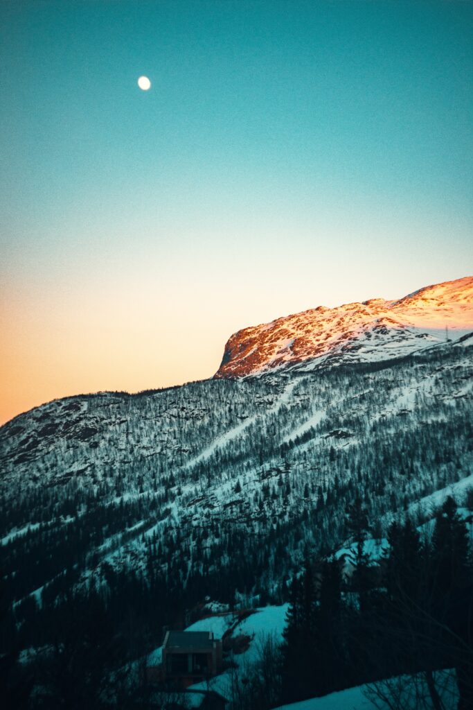 Snow-covered mountain slope at sunset with tall trees and bright moon in clear blue sky winter wallpaper
