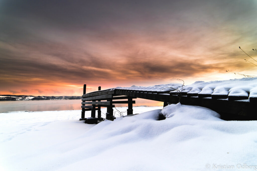 Snow-covered pier over frozen lake at vibrant orange sunset with soft clouds serene winter scene Wallpaper