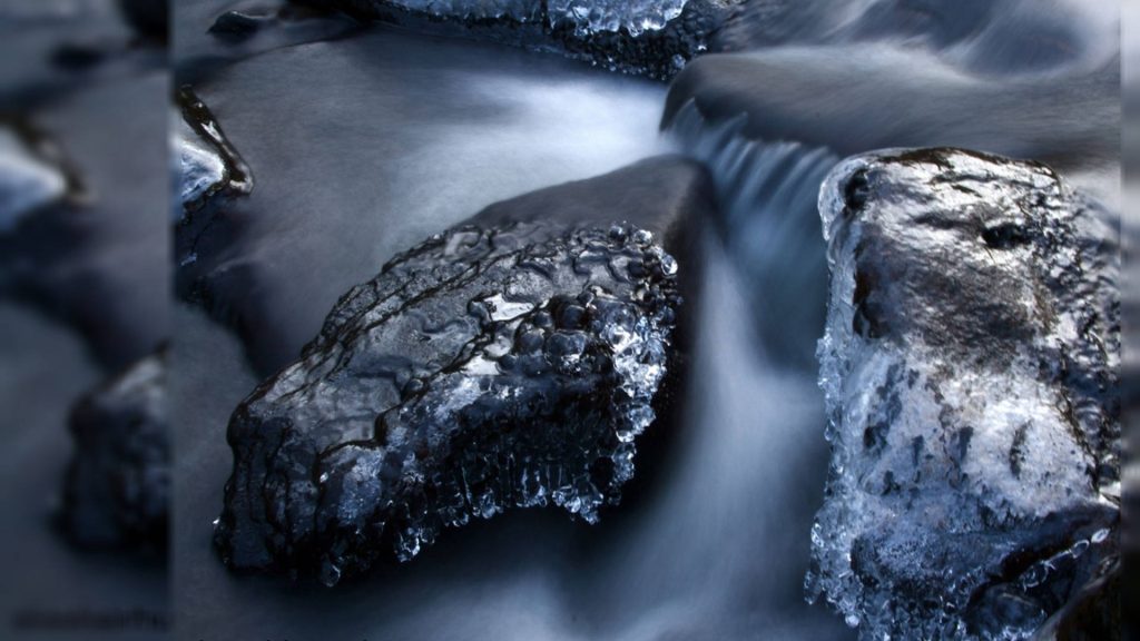 Serene long-exposure river with ice-crusted rocks and silky water in calming winter scene Wallpaper