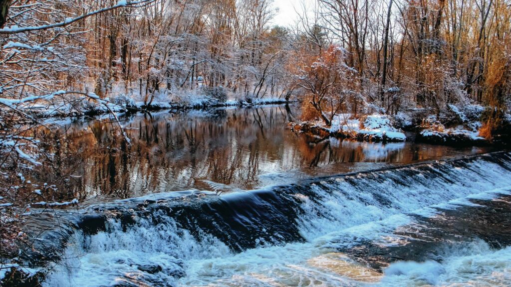 Serene winter river flowing over icy threshold with snow-dusted bare trees and sunlight reflections Wallpaper