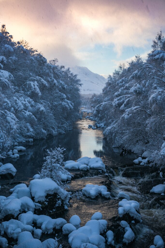 Winter river flowing through snow-covered rocks and trees with mountains and pastel sky background Wallpaper