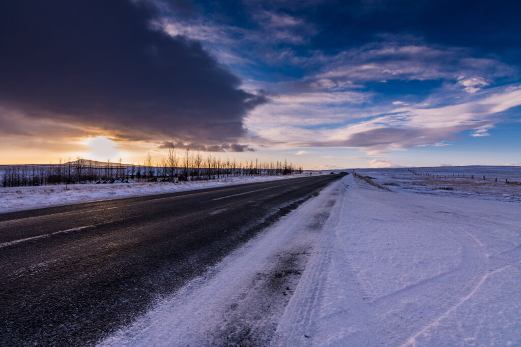 Serene winter road covered in snow under dramatic sky with soft sunlight reflecting on ice Wallpaper