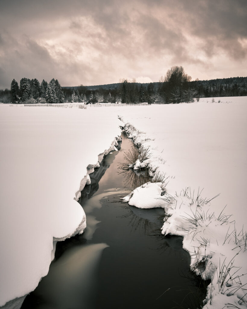 Tranquil winter landscape with narrow stream snow-covered field grasses silhouetted trees sky Wallpaper