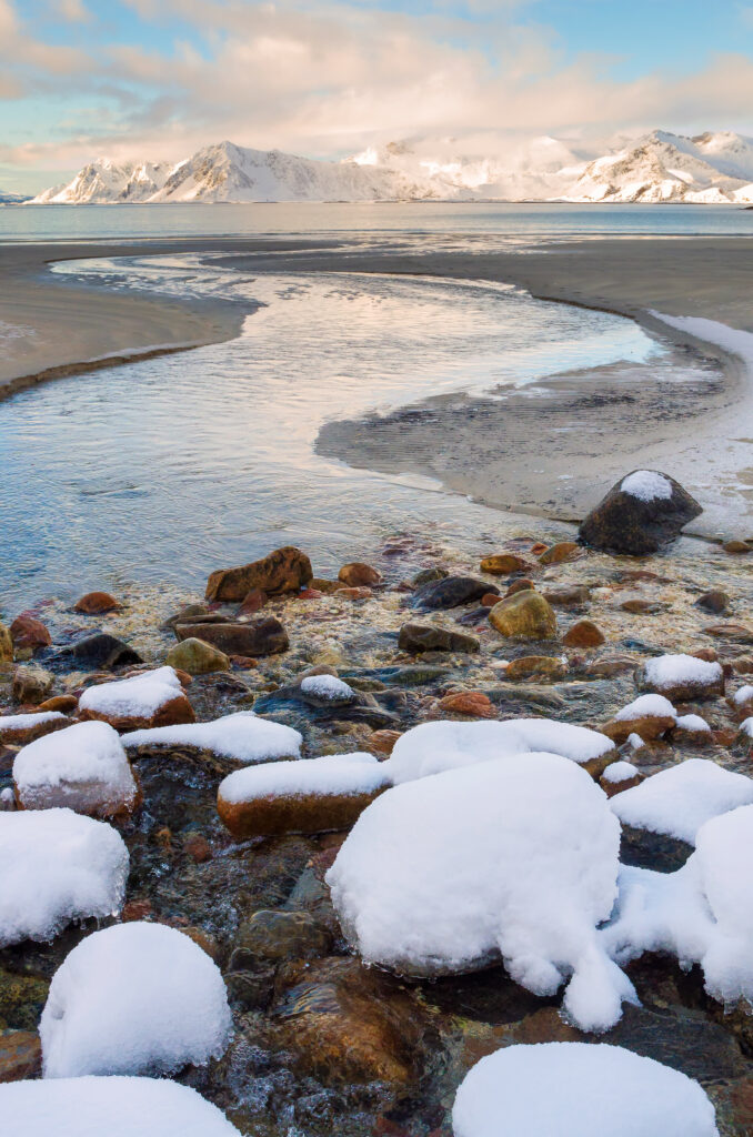 Serene winter landscape with snow-covered stream and majestic snowy mountains under blue sky Wallpaper