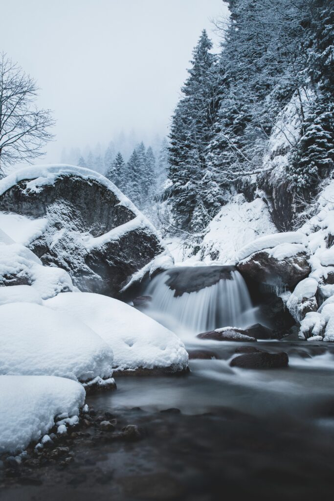 Serene winter waterfall flowing between snow-covered rocks and evergreen trees with mist Wallpaper