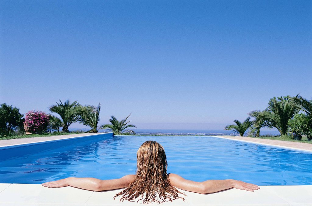Woman relaxing alone in bright azure pool with ocean view palm trees and clear summer sky Wallpaper