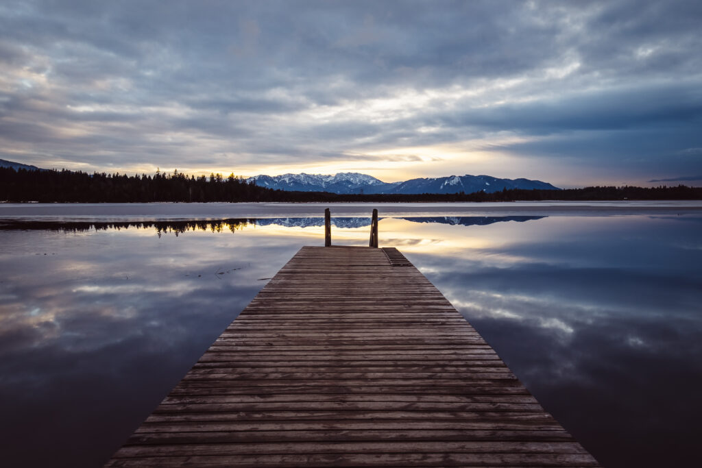 Serene wooden pier over calm lake reflecting snow-capped mountains and dramatic cloudy sky Wallpaper