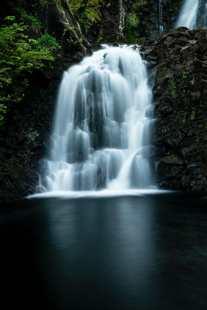 Serene waterfall flowing over rocky cliff into calm river surrounded by lush green foliage Wallpaper