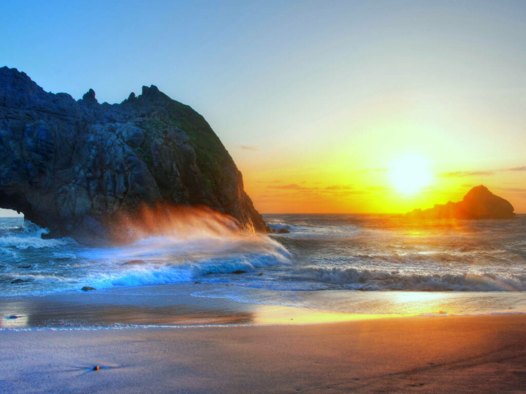 Sunset Serenade: Majestic Rock Formation Embracing Crashing Waves on Beach Desktop Wallpaper