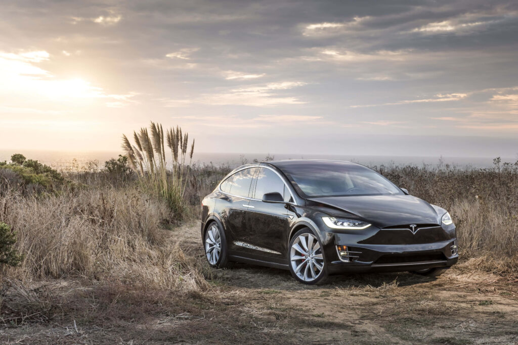 Shiny black Tesla parked on rugged dirt path with dry grass and warm sunset sky background Wallpaper