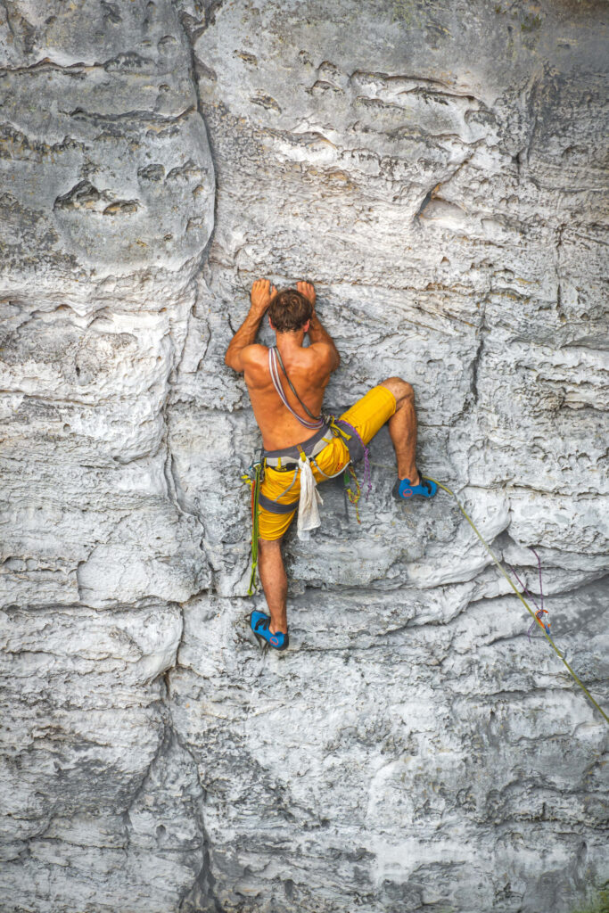 Shirtless climber in yellow shorts free climbs rugged vertical rock face showing strength Wallpaper