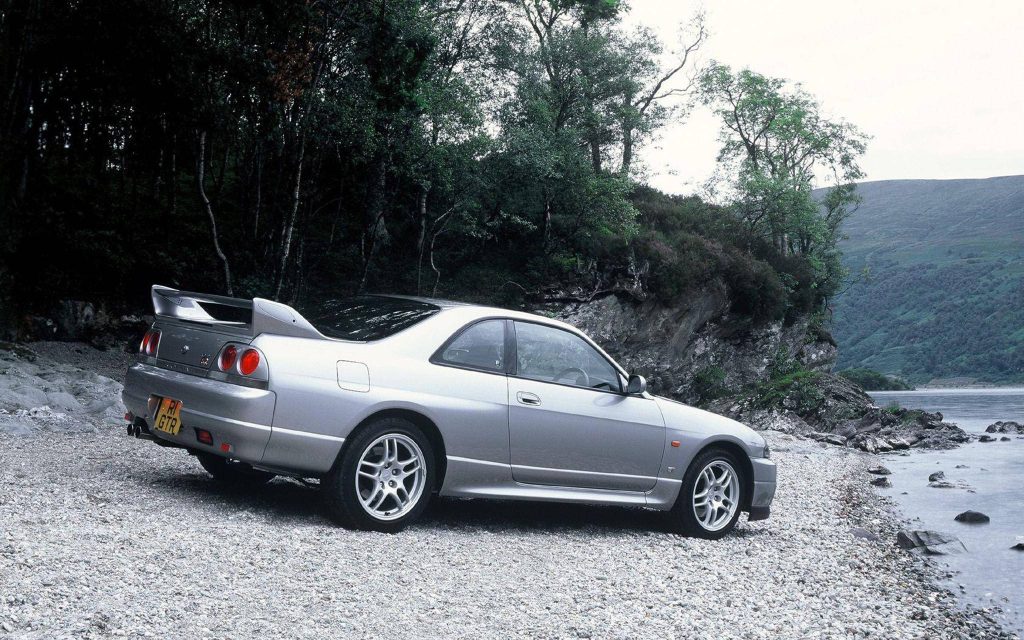 Silver Nissan Skyline GT-R R33 parked on pebbly shore with water and green woodland backdrop Wallpaper