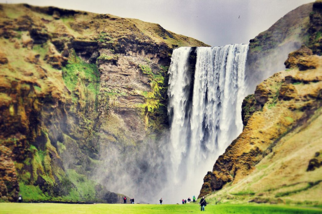 Skogafoss waterfall in Iceland cascading over cliffs with vibrant green moss and tiny hikers below Wallpaper