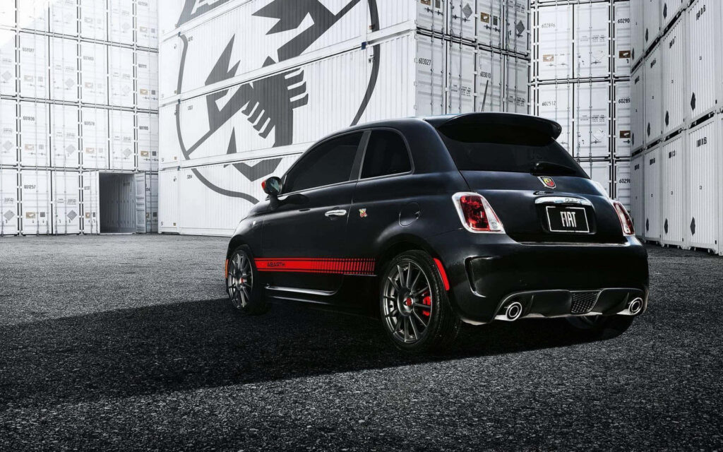 Black Abarth car with red accents and dynamic stance against industrial branded storage containers Wallpaper