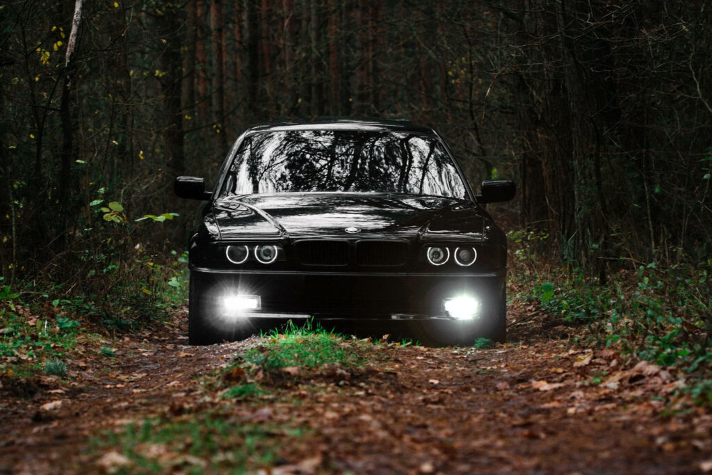Sleek black car with shining headlights on rugged dirt road in dense forest wallpaper background