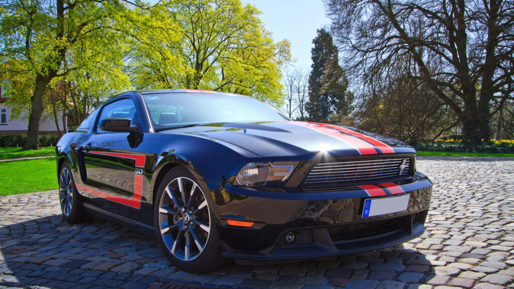 Black Mustang GT with red silver racing stripes on sunlit cobblestone driveway surrounded by trees Wallpaper