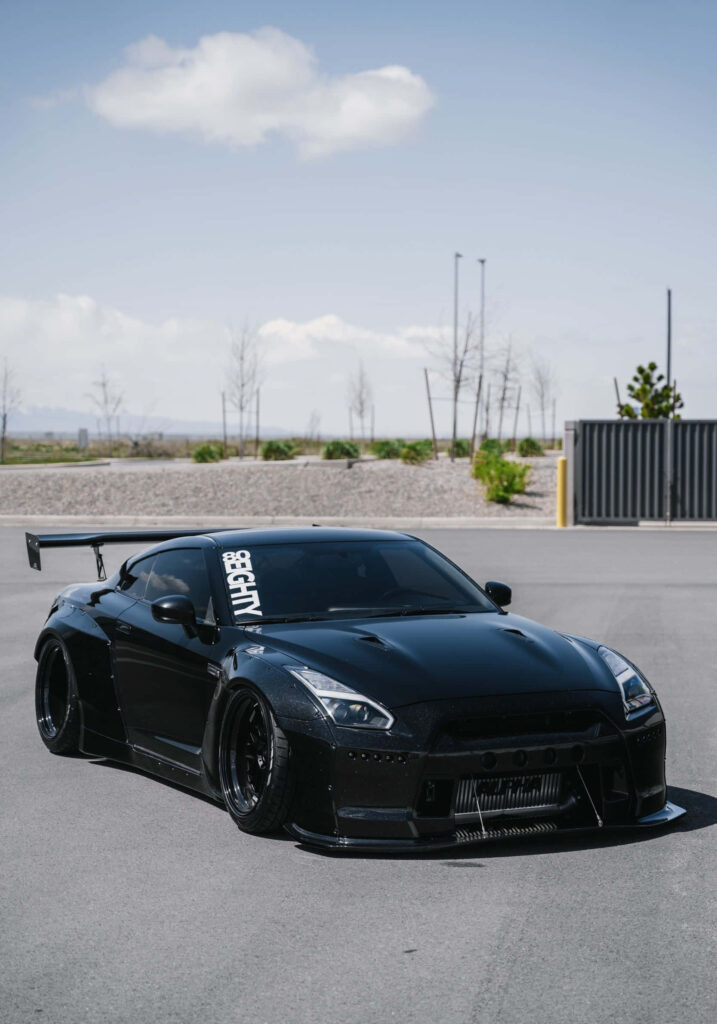 Sleek black Nissan R35 GTR with aggressive body kit and large rear wing on empty street under blue sky Wallpaper