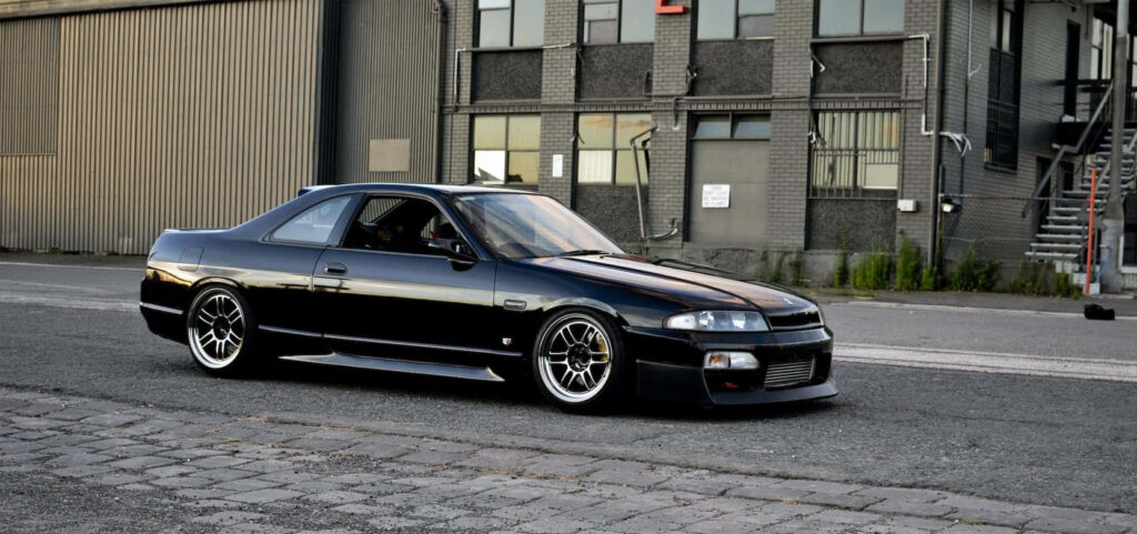 Black R33 GTR driving on city street with industrial buildings urban scenery wallpaper background