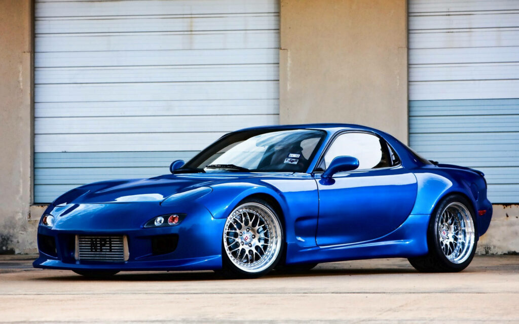 Captivating Rx7  Photo: Striking Blue Mazda Rx-7 Showcasing Silver Wheels, Skillfully Framed Against Light Blue Garage Doors and Textured Light Brown Walls Wallpaper