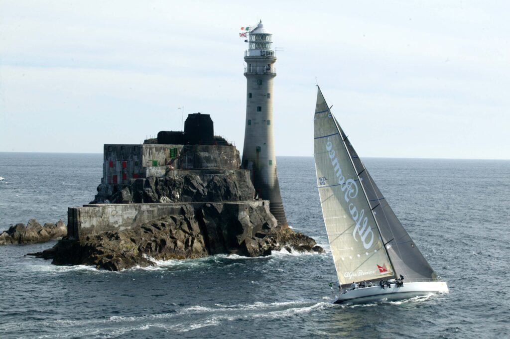 Sailing boat passes rugged rock island with weathered lighthouse and tourists under clear sky Wallpaper