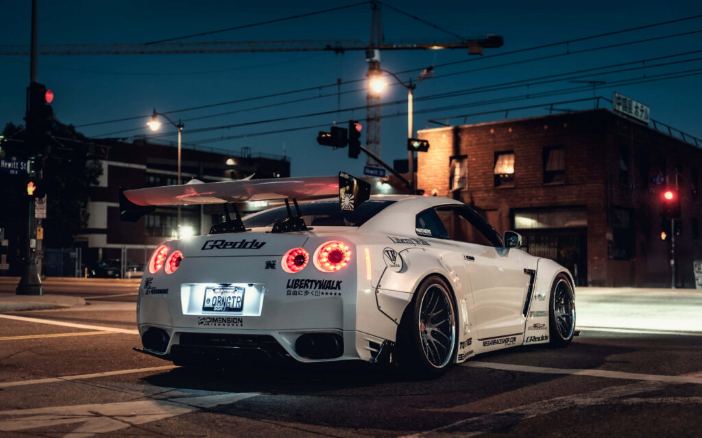 White modified sports car with rear wing and decals parked under urban city lights at night Wallpaper