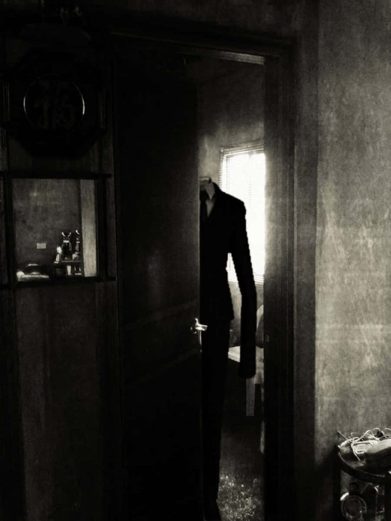 Slender Man in black suit looming in dark doorway eerie horror wallpaper creepypasta background