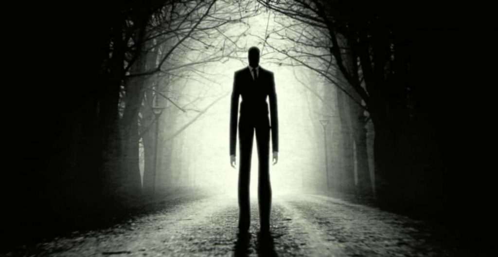 Slender Man in black suit on eerie forest path with mist and silhouetted trees horror wallpaper