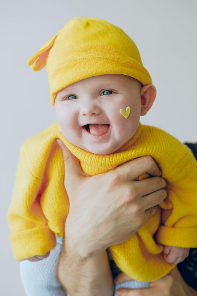 Laughing baby in bright yellow outfit and hat with heart sticker on cheek soft background wallpaper
