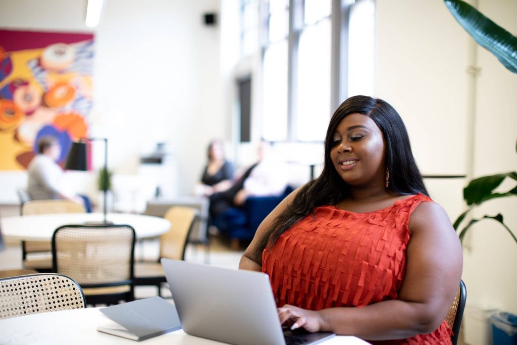 Plus-size Black woman in orange top working on laptop in bright modern office with mural background Wallpaper