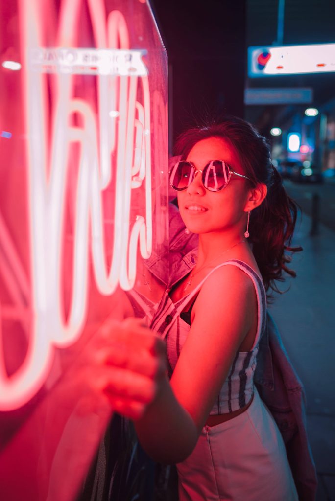 Smiling woman in round sunglasses beside vibrant neon red sign at night cinematic urban background Wallpaper