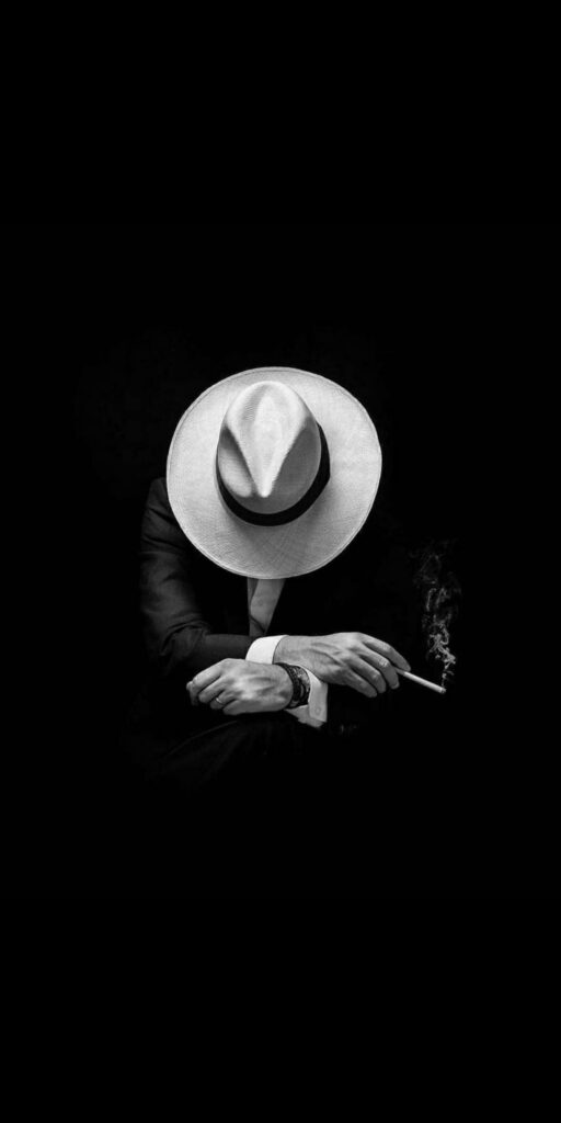 Mysterious man in suit and fedora smoking in high contrast black and white noir wallpaper background